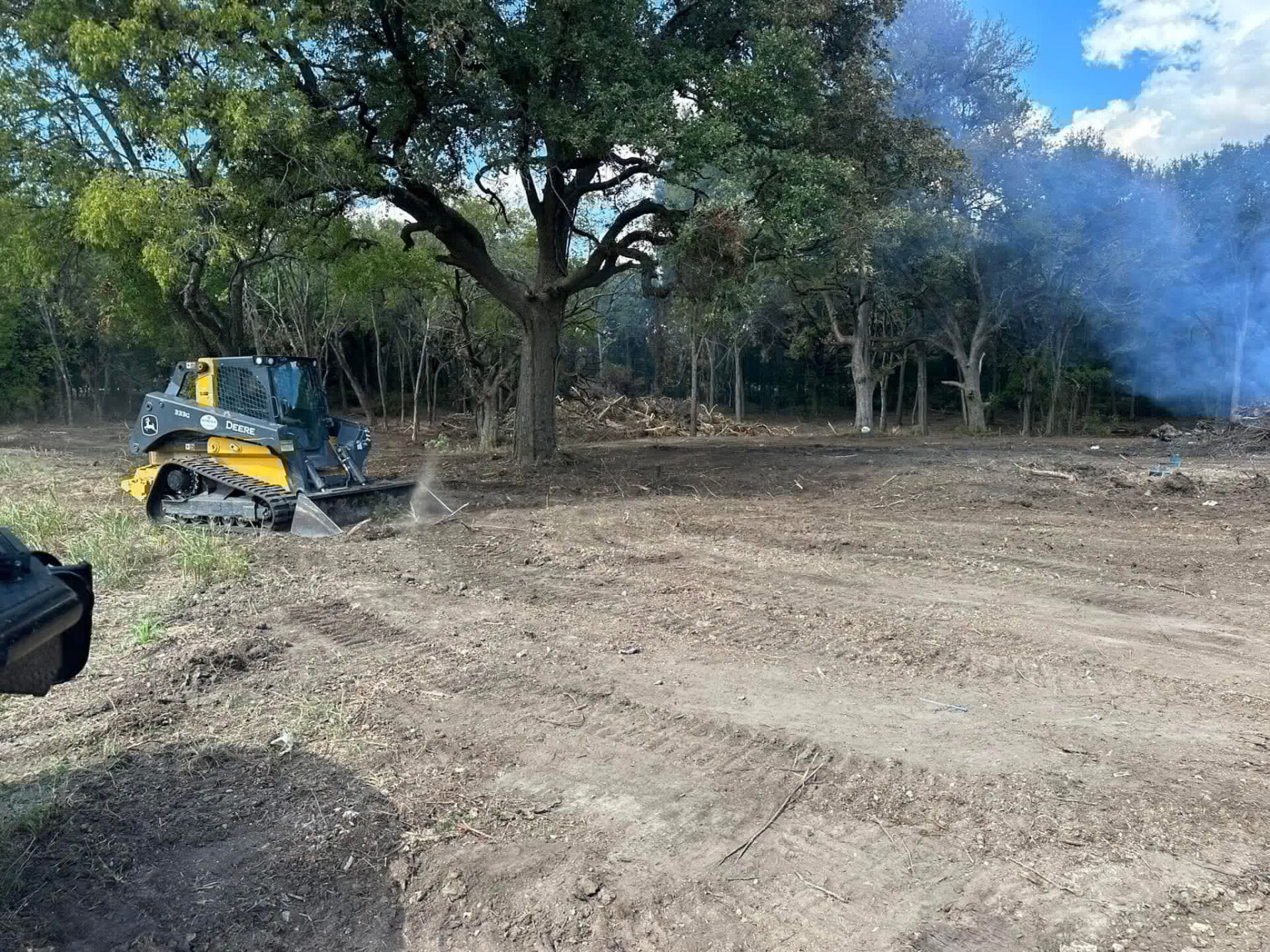 Deere compact track loader clearing brush beneath large oak trees